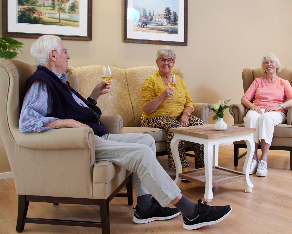 Bewohner des Gemeinschafts-Wohnens sitzen im einer Sitzgruppe und stoßen mit Wein an