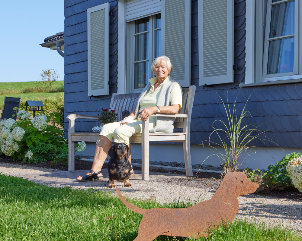 Bewohnerin sitzt vor einem idyllischen Landhaus im Landhaus-Wohnen auf einer Bank
